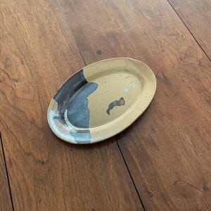 May include: An oval ceramic dish with a speckled tan base glaze. One side of the dish features a smooth, dark charcoal-grey glaze that overlaps the tan surface in an irregular, flowing pattern. The dish has a slightly raised rim and a glossy finish.