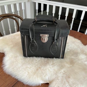 May include: A black structured vanity case with a top handle, a silver-toned metal latch closure, and two vertical side zippers. The case features a small silver-toned key emblem attached to the top lid, providing a secure storage solution.