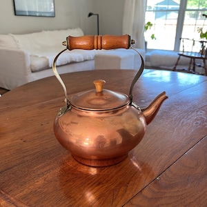 May include: A copper tea kettle with a rounded body, a short spout, and a lid featuring a small wooden knob. The kettle has a brass-toned metal handle with a cylindrical wooden grip centered at the top, providing a contrast to the metallic finish.