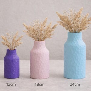 Pode incluir: Três vasos decorativos em roxo, rosa e azul, cada um com uma superfície texturizada e cheios de flores secas. Os vasos têm 12 cm, 18 cm e 24 cm de altura, respetivamente. Os vasos estão dispostos da esquerda para a direita.