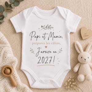 Peut inclure: Body de bébé blanc avec l'inscription "Papi et Mamie, préparez les câlins, J'arrive en 2027!" en écriture noire et marron. Un cœur en bois, un lapin en peluche et un hochet en bois sont également présents.