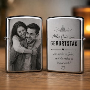 Puede incluir: Dos encendedores de metal plateado. El encendedor de la izquierda presenta una fotografía en blanco y negro de una pareja sonriente abrazada. El de la derecha muestra el texto en alemán Alles Gute zum GEBURTSTAG, un icono de pastel y un corazón.