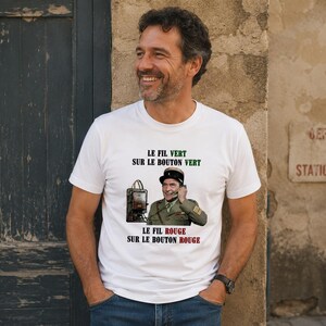Peut inclure: Un t-shirt blanc à manches courtes avec une impression graphique représentant un officier militaire en uniforme tenant un combiné téléphonique. Le texte indique : LE FIL VERT SUR LE BOUTON VERT et LE FIL ROUGE SUR LE BOUTON ROUGE.