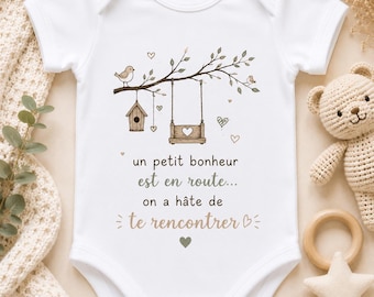 May include: A white short-sleeve baby bodysuit featuring a graphic illustration of a tree branch with a birdhouse, a swing, and small birds. The text reads: un petit bonheur est en route... on a hâte de te rencontrer, accented with hearts.