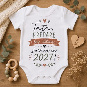 Peut inclure: Un body pour bébé blanc à manches courtes avec le texte noir et terracotta "Tata, PRÉPARE les câlins, j'arrive en 2027!" accompagné de motifs de cœurs et de feuilles. Le vêtement possède une encolure ronde et trois boutons-pression en bas.