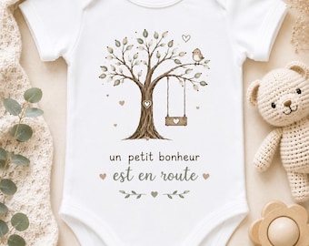 May include: A white short-sleeved baby bodysuit featuring a brown tree illustration with a swing, a small bird, and hearts. The text "un petit bonheur est en route" is printed in brown below the tree, accompanied by small heart and leaf accents.