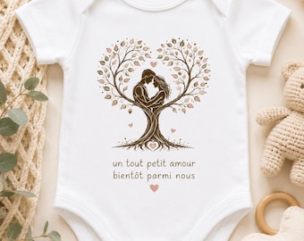 May include: A white short-sleeved baby bodysuit featuring a brown graphic of two people embracing within a heart-shaped tree. Below the illustration, the text reads: un tout petit amour bientôt parmi nous, accompanied by small pink heart accents.