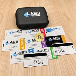 Puede incluir: Un estuche negro con cremallera con el logotipo "LABS TERRAGROUP", un Bitcoin dorado y varias tarjetas de acceso blancas con etiquetas de colores. Las tarjetas tienen texto y el mismo logotipo. El fondo es una superficie de madera clara.