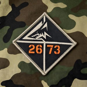 May include: A diamond-shaped fabric patch with a dark navy blue background and a light beige stitched border. The design features a white stylized shark graphic above the orange embroidered numbers 26 and 73, separated by a thin white line.