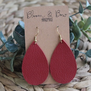 Puede incluir: Un par de pendientes de cuero rojo en forma de lágrima con ganchos dorados. Los pendientes están hechos a mano y se muestran en una tarjeta con el texto "Bloom & Bow Handmade".
