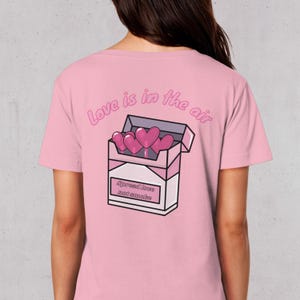 Könnte beinhalten: Hellrosa T-Shirt mit einem Aufdruck auf der Rückseite, der eine Zigarettenschachtel mit rosa Herzen und dem Text "Love is in the air" und "Spread love not smoke" zeigt.