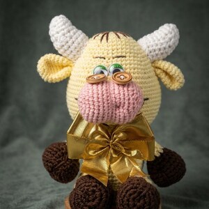 May include: A handmade crochet cow doll with a yellow body, white horns, and brown hooves. The cow has a pink snout, button eyes, and a gold ribbon bow. The doll is on a wooden base with a "HANDMADE ARTISTRY" label.