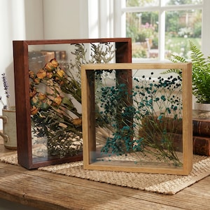 May include: Two wooden shadow boxes with glass fronts. One is dark brown and contains dried yellow roses. The other is light brown and holds blue baby's breath. Both are displayed on a wooden table with a woven mat.
