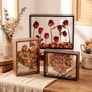 May include: Several wooden framed displays with dried roses and petals. One frame contains a bouquet tied with a ribbon. Another frame features red roses with petals and pearls. The frames are on a wooden table.