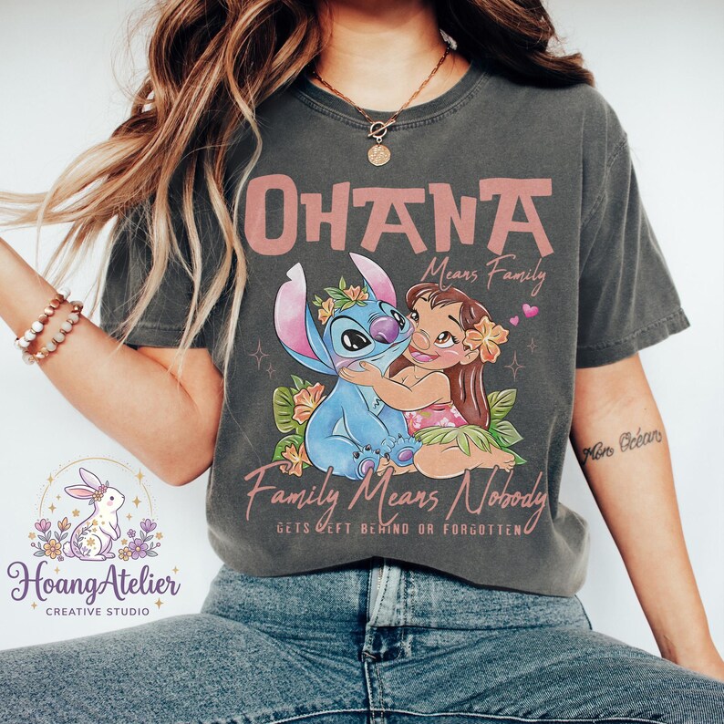 Op de afbeelding: Donkergrijs t-shirt met een afbeelding van Stitch en Lilo, en de tekst "OHANA Means Family" en "Family Means Nobody Gets Left Behind or Forgotten." Het ontwerp bevat bloemmotieven en harten.