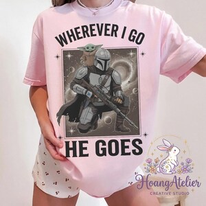 May include: A light pink t-shirt featuring a graphic of The Mandalorian and Grogu with the text "WHEREVER I GO HE GOES". The design includes a space background and stars.