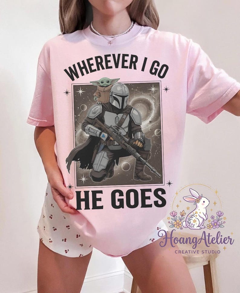 May include: A light pink t-shirt featuring a graphic of The Mandalorian and Grogu with the text "WHEREVER I GO HE GOES". The design includes a space background and stars.