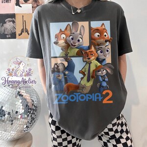 K&ouml;nnte beinhalten: Dunkelgraues T-Shirt mit einer farbenfrohen Grafik von Charakteren aus dem Animationsfilm "Zoomania". Das Design enth&auml;lt den Text "Zoomania 2" in Blau und Orange. Ein l&auml;ssiges Kleidungsst&uuml;ck.