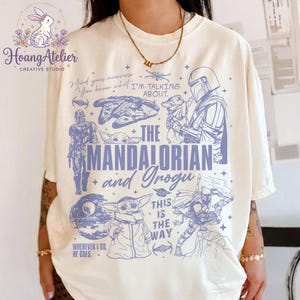 Puede incluir: Camiseta color crema con un gr&aacute;fico azul de personajes y elementos de la serie The Mandalorian. El dise&ntilde;o incluye el texto "The Mandalorian and Grogu" y otras frases. La camiseta tiene cuello redondo y mangas cortas.