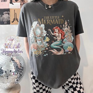 May include: Dark gray t-shirt featuring a colorful illustration of Ariel from "The Little Mermaid." The design includes Ariel, a castle, a treasure chest, and the text "The Little Mermaid" in gold lettering. The shirt is a comfortable, casual style.