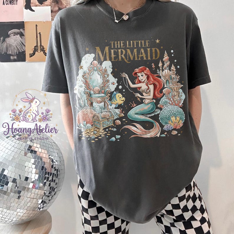 May include: Dark gray t-shirt featuring a colorful illustration of Ariel from "The Little Mermaid." The design includes Ariel, a castle, a treasure chest, and the text "The Little Mermaid" in gold lettering. The shirt is a comfortable, casual style.