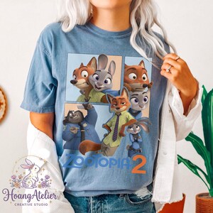 K&ouml;nnte beinhalten: Hellblaues T-Shirt mit einem Aufdruck von Charakteren aus dem Animationsfilm Zootopia. Das Design zeigt einen Fuchs, ein Kaninchen und andere Charaktere mit dem Text "Zootopia 2".