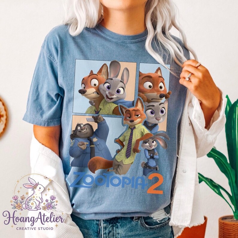 K&ouml;nnte beinhalten: Hellblaues T-Shirt mit einem Aufdruck von Charakteren aus dem Animationsfilm Zootopia. Das Design zeigt einen Fuchs, ein Kaninchen und andere Charaktere mit dem Text "Zootopia 2".