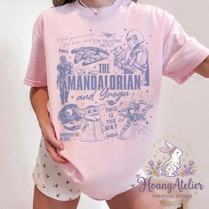 Puede incluir: Camiseta rosa claro con un dise&ntilde;o gr&aacute;fico que presenta el texto "The Mandalorian and Grogu". El dise&ntilde;o incluye ilustraciones de personajes y elementos de la serie Star Wars. La camiseta tiene mangas cortas.