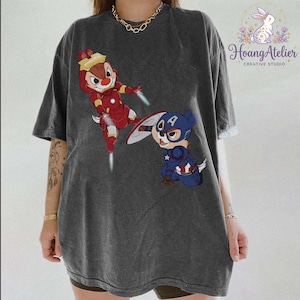 May include: Dark gray t-shirt featuring cartoon characters Chip and Dale dressed as Iron Man and Captain America. The Iron Man Chipmunk is in mid-air with a red and gold suit. The Captain America Chipmunk is in a blue suit with a shield.