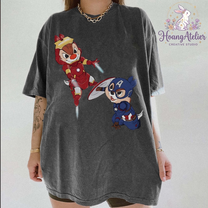 May include: Dark gray t-shirt featuring cartoon characters Chip and Dale dressed as Iron Man and Captain America. The Iron Man Chipmunk is in mid-air with a red and gold suit. The Captain America Chipmunk is in a blue suit with a shield.
