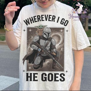 May include: Cream-colored t-shirt featuring a graphic of The Mandalorian and Grogu. The design includes the text "WHEREVER I GO HE GOES" in bold, black letters. The image depicts the Mandalorian kneeling with a rifle, and Grogu is perched on his shoulder.