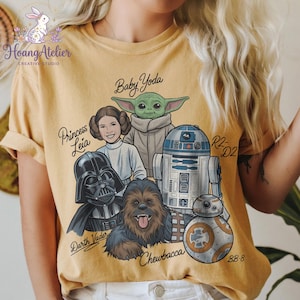 May include: Mustard yellow t-shirt featuring a graphic of Star Wars characters: Darth Vader, Princess Leia, Baby Yoda, Chewbacca, R2-D2, and BB-8. The shirt has the text "Hoang Atelier CREATIVE STUDIO" and character names. The design is printed on a soft, comfortable tee.