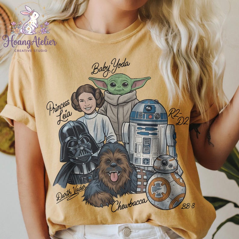 May include: Mustard yellow t-shirt featuring a graphic of Star Wars characters: Darth Vader, Princess Leia, Baby Yoda, Chewbacca, R2-D2, and BB-8. The shirt has the text "Hoang Atelier CREATIVE STUDIO" and character names. The design is printed on a soft, comfortable tee.