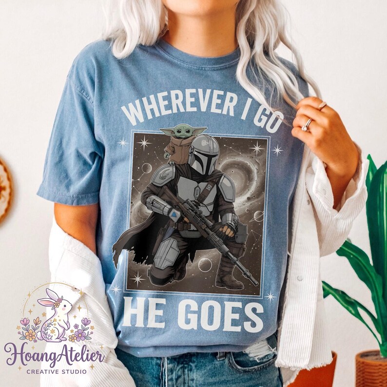 May include: Blue t-shirt with the text "WHEREVER I GO HE GOES" and a graphic of The Mandalorian and Grogu. The design features a space background and is framed in a white rectangle. The shirt is a light blue color.