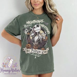 May include: Olive green t-shirt featuring a circular graphic from "The Nightmare Before Christmas." The design includes Jack Skellington, Sally, and pumpkins, with the text "Nightmare Before Christmas" and "The Pumpkin King."