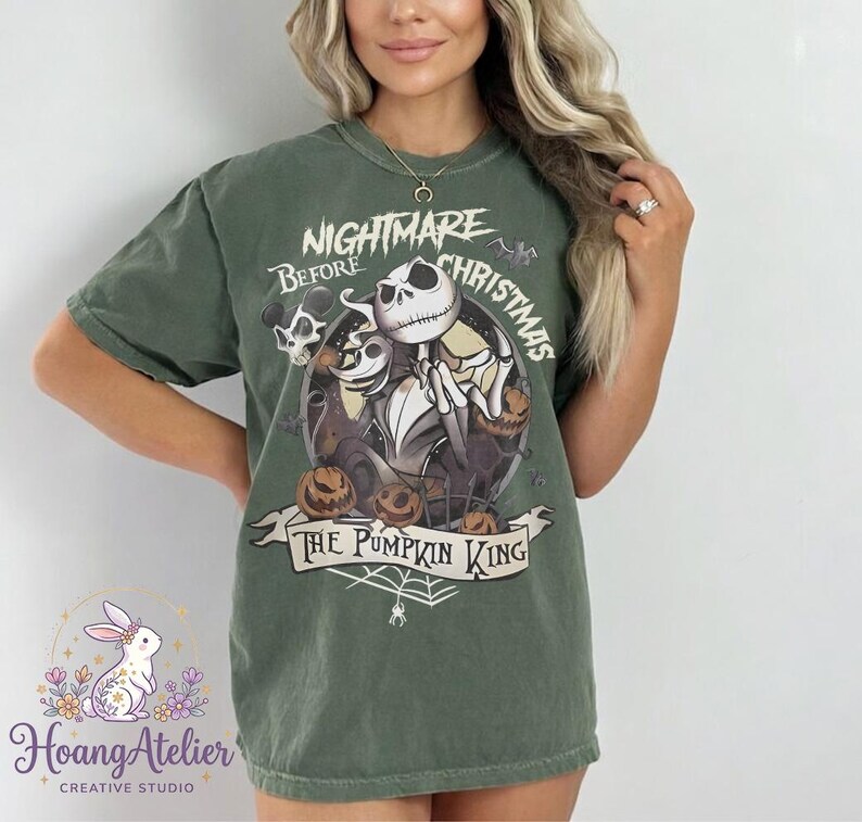 May include: Olive green t-shirt featuring a circular graphic from "The Nightmare Before Christmas." The design includes Jack Skellington, Sally, and pumpkins, with the text "Nightmare Before Christmas" and "The Pumpkin King."