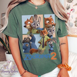 K&ouml;nnte beinhalten: Olivgr&uuml;nes T-Shirt mit einem farbenfrohen Grafikdesign von Charakteren aus dem Film "Zootopia 2". Das Design zeigt einen Fuchs, ein Kaninchen und andere Charaktere. Der Text "Zootopia 2" ist in Blau und Orange dargestellt.