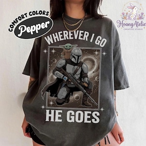 May include: Pepper-colored t-shirt featuring a graphic of The Mandalorian and Grogu with the text "WHEREVER I GO HE GOES." The design includes a space background and star details. The shirt has a relaxed fit.