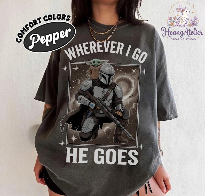 May include: Pepper-colored t-shirt featuring a graphic of The Mandalorian and Grogu with the text "WHEREVER I GO HE GOES." The design includes a space background and star details. The shirt has a relaxed fit.