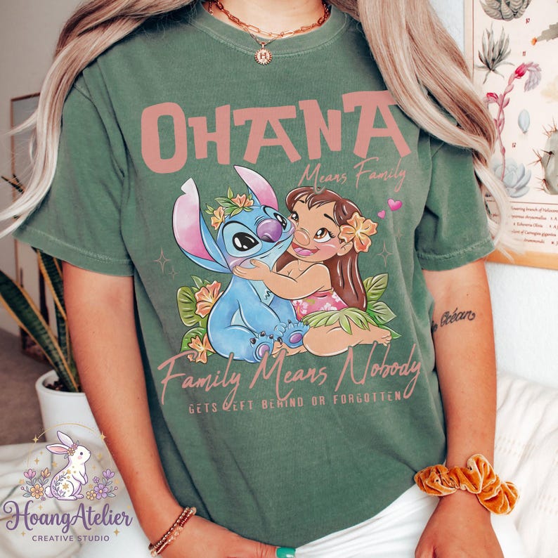 Op de afbeelding: Groen t-shirt met de roze tekst "OHANA Means Family". De afbeelding toont Stitch en Lilo die elkaar omhelzen, omringd door bloemen. De tekst "Family Means Nobody Gets Left Behind Or Forgotten" staat er ook op.