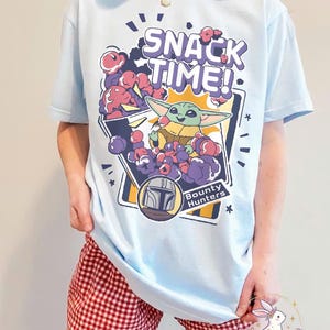 Puede incluir: Camiseta azul claro con un dibujo animado de Grogu de The Mandalorian, rodeado de snacks morados y rojos, con el texto "SNACK TIME!". La camiseta tambi&eacute;n incluye el texto "Bounty Hunters" y un gr&aacute;fico de casco.