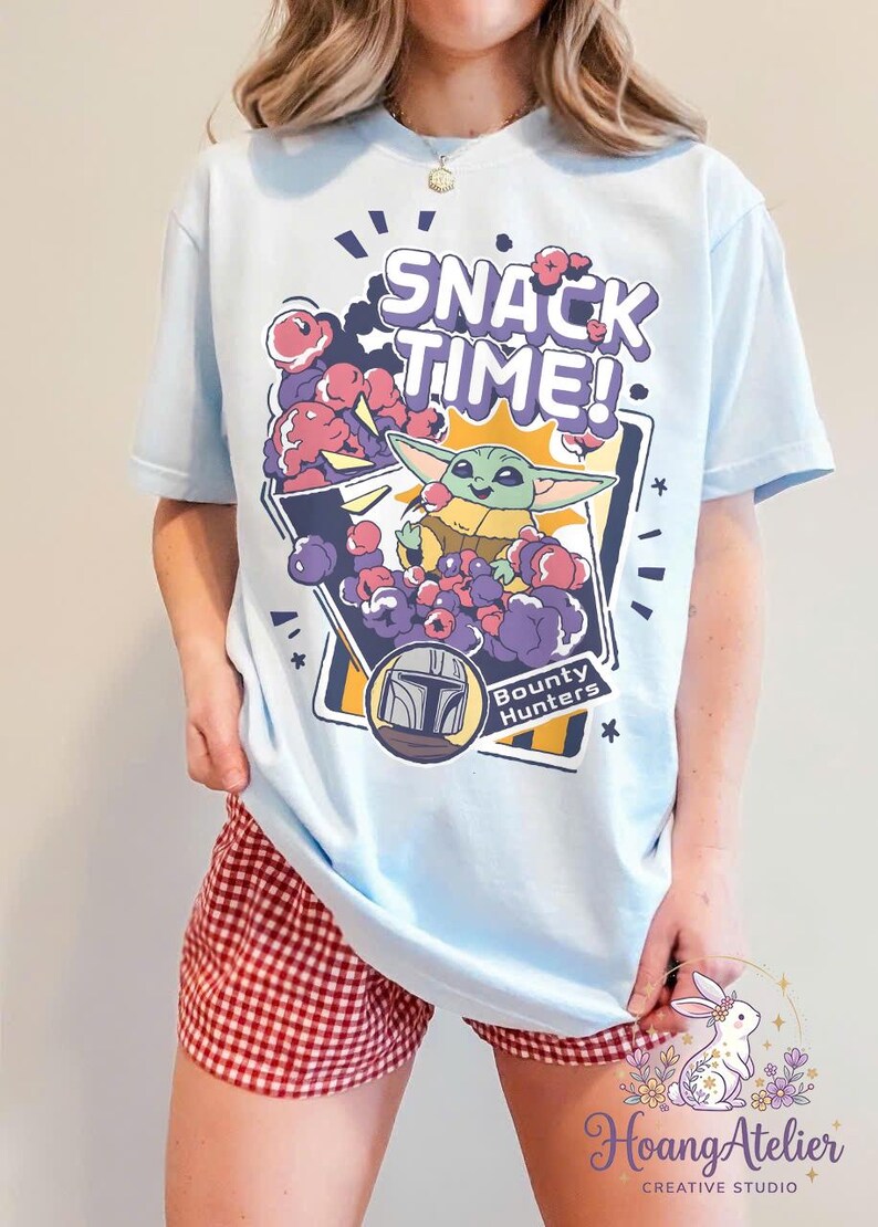 Puede incluir: Camiseta azul claro con un dibujo animado de Grogu de The Mandalorian, rodeado de snacks morados y rojos, con el texto "SNACK TIME!". La camiseta tambi&eacute;n incluye el texto "Bounty Hunters" y un gr&aacute;fico de casco.