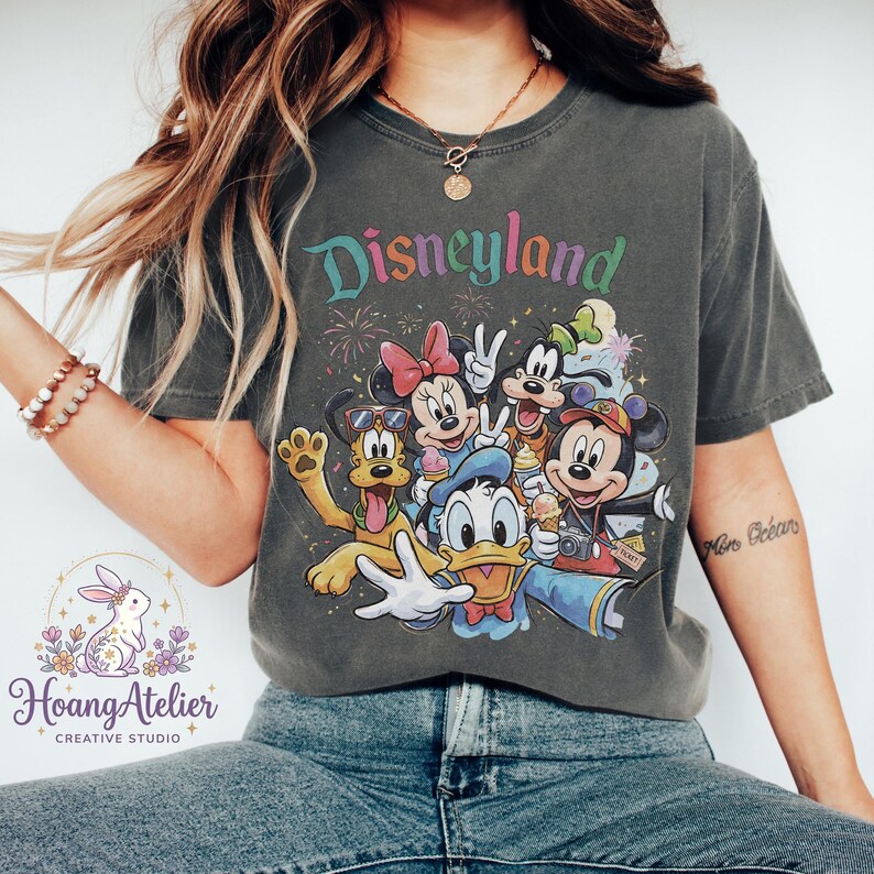 May include: Dark gray t-shirt featuring the word "Disneyland" in colorful text above a graphic of Disney characters including Mickey Mouse, Minnie Mouse, Donald Duck, and Pluto. The shirt has a vintage, worn look.
