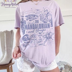 Puede incluir: Camiseta lila claro con ilustraciones de "The Mandalorian", incluyendo personajes y frases. El dise&ntilde;o incluye el texto "The Mandalorian and Grogu". La camiseta se usa con pantalones cortos blancos.