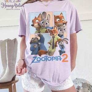 K&ouml;nnte beinhalten: Helllila T-Shirt mit einem Cartoon-Motiv von Charakteren aus dem Film "Zootopia 2". Das Design zeigt einen Fuchs, ein Kaninchen und andere Charaktere. Der Text "Zootopia 2" ist in Blau und Orange gedruckt.
