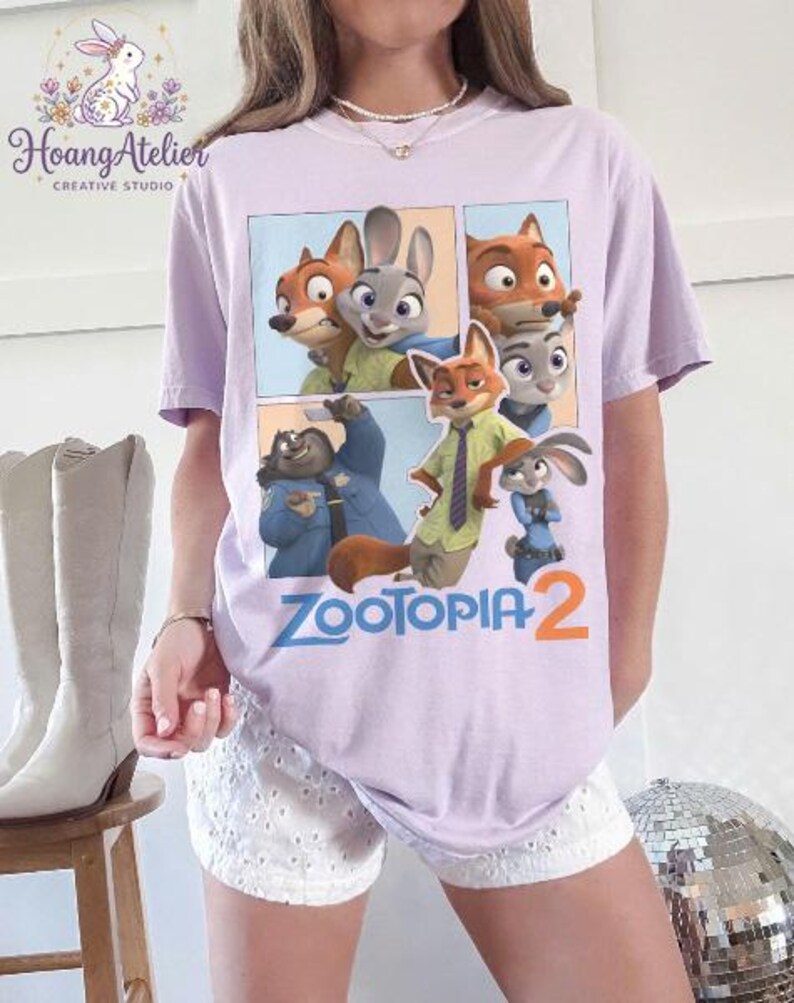K&ouml;nnte beinhalten: Helllila T-Shirt mit einem Cartoon-Motiv von Charakteren aus dem Film "Zootopia 2". Das Design zeigt einen Fuchs, ein Kaninchen und andere Charaktere. Der Text "Zootopia 2" ist in Blau und Orange gedruckt.