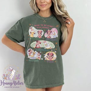 May include: Olive green t-shirt with a graphic of Minnie Mouse and friends, and the text "HOW TO BE KIND WITH MINNIE!". The design includes illustrations of Minnie, Daisy, and other characters. The shirt also has the Hoang Atelier logo.