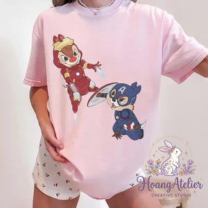 May include: Light pink t-shirt featuring cartoon characters Chip and Dale dressed as Iron Man and Captain America. The Iron Man Chipmunk is in a red and gold suit, while Captain America Dale is in a blue and red costume. The shirt has the "Hoang Atelier" logo.