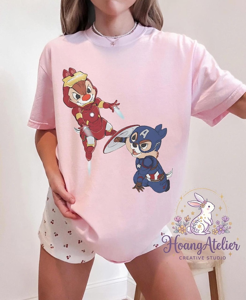 May include: Light pink t-shirt featuring cartoon characters Chip and Dale dressed as Iron Man and Captain America. The Iron Man Chipmunk is in a red and gold suit, while Captain America Dale is in a blue and red costume. The shirt has the "Hoang Atelier" logo.