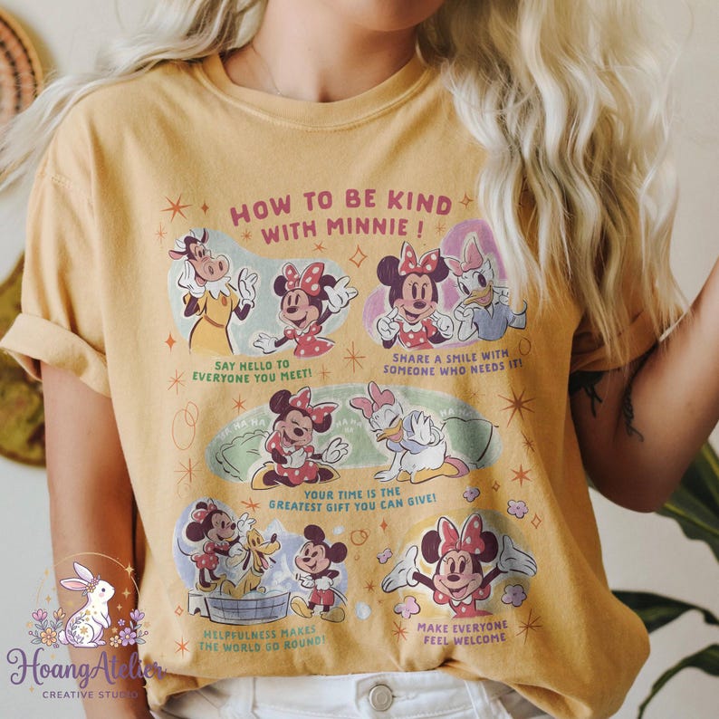 May include: Mustard yellow t-shirt with a graphic of Minnie Mouse and friends, including text that reads "HOW TO BE KIND WITH MINNIE!". The graphic also features kindness tips. The sleeves of the shirt are rolled up.